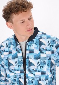 Giacca bomber blu con un collage di onde e scene di spiaggia. Realizzata in tessuto leggero con colletto nero e chiusura a zip.