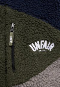 Gros plan d'une veste en polaire avec une poche zippée verte et un texte brodé blanc « UNFAIR Athletics » sur tissu vert.