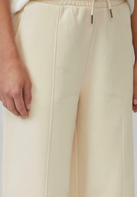 Nahaufnahme von hellbeigen Hosen mit vertikalen Nähten und weißem Kordelzug, mit einer ruhenden Hand neben dem Bein.