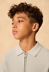 Light gray zip-up jacket with a textured fabric, collared design, and silver zipper. Model has curly hair and is posed against a neutral background.
