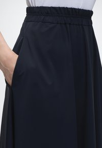 Personne portant une jupe noire avec une ceinture élastique et la main placée dans la poche latérale sur un fond blanc.