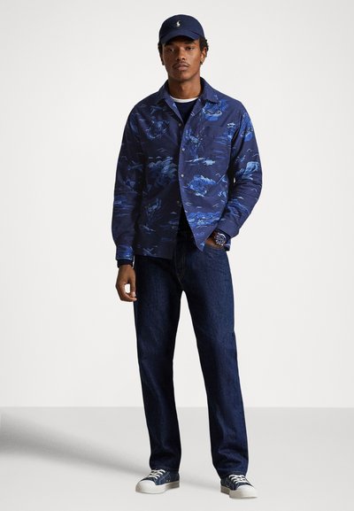 Μπλε μπουφάν με μοτίβα και μακριά μανίκια, κλείσιμο με κουμπιά και αντίθετο μπλε τζιν παντελόνι. Το μοντέλο φοράει ναυτικό καπέλο και λευκά αθλητικά παπούτσια.