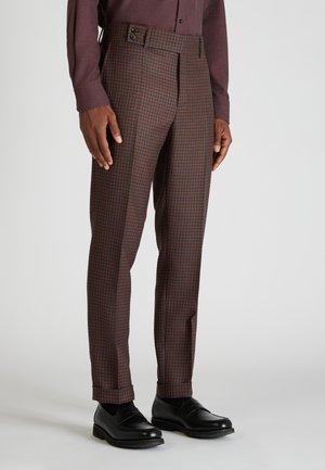 Homme portant un pantalon sur mesure à carreaux marron et bleu, une chemise à manches longues bordeaux et des mocassins en cuir noir sur un fond uni.