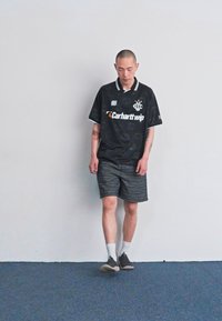 Fekete póló fehér gallérral és logóval, sötét mintás rövidnadrággal és szürke sportcipővel párosítva. Semleges háttér előtt áll.