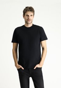 Camiseta negra de manga corta hecha de una tela suave con cuello redondo, llevada con jeans negros, manos en los bolsillos, fondo neutro.