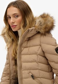 Beige pufferjakke med hætte foret med pels. Den har vandret quiltning, lynlås lukning og en sidelomme med lynlås. Tekstureret stof.