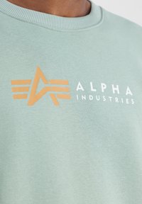 Svijetlozeleni dukserica s rebrastim ovratnikom. Sadrži narančasti logo s geometrijskim oblicima i natpisom "ALPHA INDUSTRIES" u bijeloj boji.