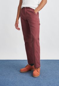 Homemem usando calças de perna reta na cor ferrugem e sapatos de couro laranja-escuro combinando, em pé sobre um tapete azul com uma mão no bolso.