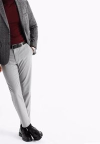 Homme portant un blazer gris à carreaux, un col roulé bordeaux, un pantalon gris clair, une ceinture noire et des chaussures de ville noires, debout les jambes croisées.