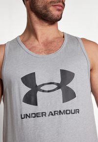 Chemise sans manches grise en mélange de coton, présentant un grand logo Under Armour noir et du texte à l'avant. Texture lisse et coupe décontractée.