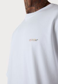 White cotton t-shirt with short sleeves, featuring a small multicolored embroidered logo "ESTEEM" on the left chest area. Smooth texture.