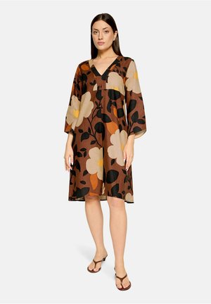Femme portant une robe marron mi-longue avec un grand imprimé floral beige, noir et orange, col en V, manches larges et sandales marron.