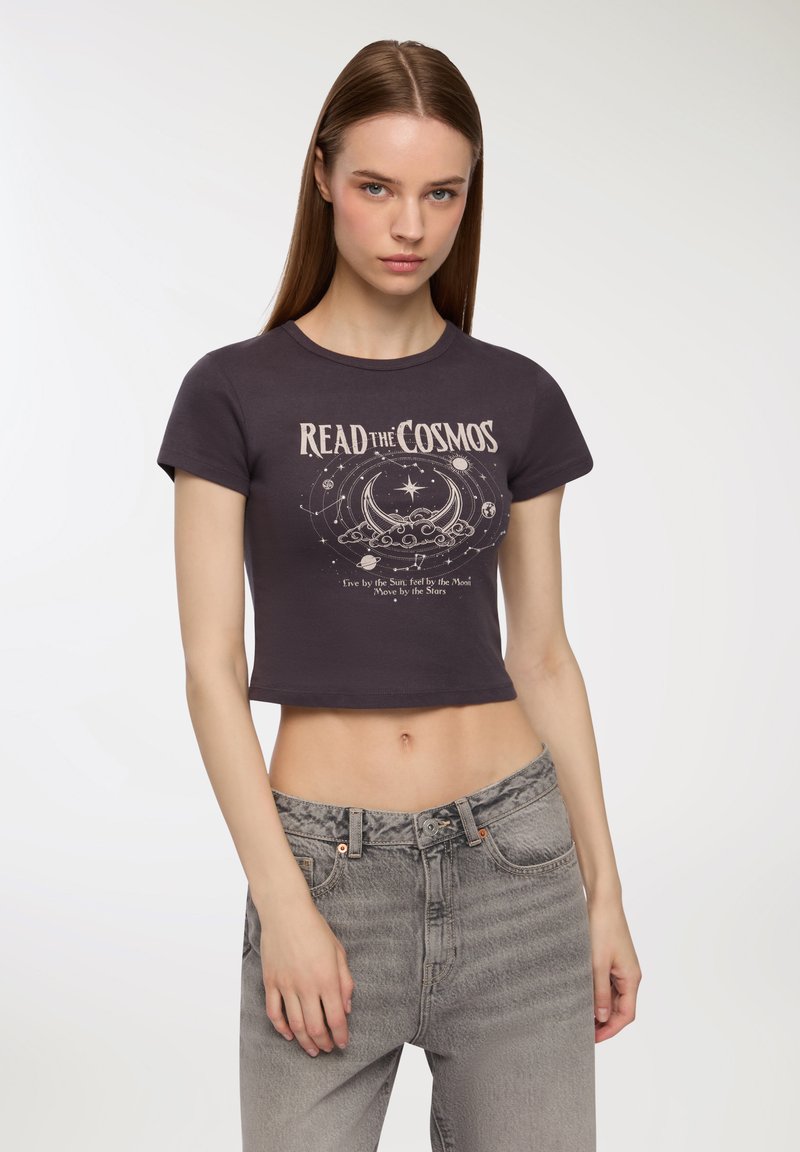 T-shirt corto grigio scuro con maniche corte, caratterizzato da una grafica celestiale bianca e la scritta "LEGGI IL COSMO". Indossato con jeans chiari a vita alta.