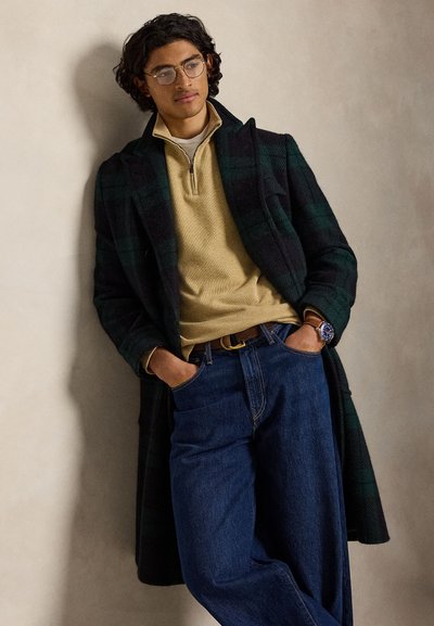 Jeune homme portant des lunettes, un pull beige à demi-fermeture éclair, un manteau sombre à carreaux, un jean large bleu et une montre, appuyé contre un mur beige, les mains dans les poches.