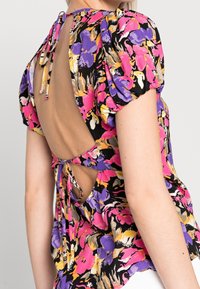 Blouse fleurie présentant un mélange de tons roses, violets et jaunes. Manches bouffantes, dos ouvert avec fermeture à nouer, et tissu léger.