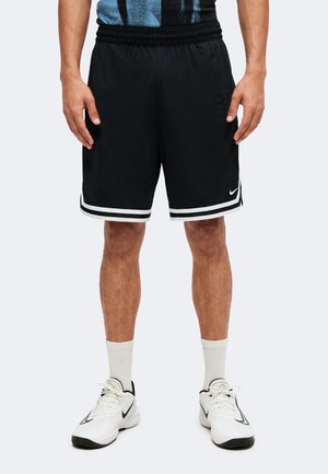 Homme portant un short de sport noir avec des bandes blanches et des baskets Nike blanches avec des chaussettes blanches, se tenant devant un fond uni.