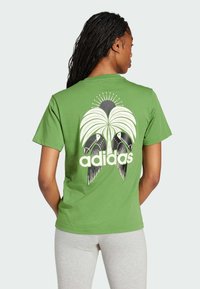 Green cotton t-shirt featuring a large graphic of a palm tree, black birds, and the "adidas" logo in white, short sleeves, relaxed fit.