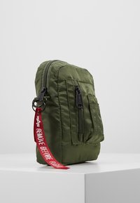 Zielona torba nylonowa w prostokątnym kształcie, z przednią kieszenią, czarnymi zamkami błyskawicznymi oraz czerwoną metką z napisem "USUNĄĆ PRZED LOTEM."