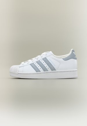 Λευκό αθλητικό παπούτσι Adidas Superstar με ανοιχτόγριμες ρίγες, κελυφωτό δάχτυλο και κείμενο «SUPERSTAR» στο πλάι, εμφανιζόμενο σε δεξιά πλευρική όψη μπροστά σε ουδέτερο φόντο.