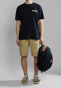 T-shirt nera in cotone con logo frontale, pantaloni cargo khaki, sneaker verdi e beige, e uno zaino nero con logo e tasca in rete.