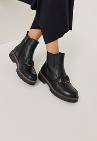 Schwarze Leder-Stiefeletten mit glänzendem Schnallenakzent, strukturiertem Fußbett und kontrastierenden, tanfarbenen Nähten. Reißverschluss an den Seiten.