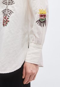 Bílá bavlněná košile s barevným květinovým vyšíváním na rukávu a hrudi, s nařaseným manžetem a zaobleným lemem.