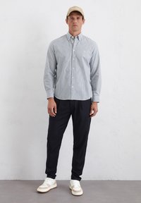 Blue and white striped button-up shirt, long sleeves, paired with dark trousers and white sneakers featuring beige accents. Beige cap on head.