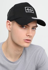 Black baseball cap with mesh back and a flat brim. Features a white "New Era" logo patch. Smooth fabric texture. Close-up view on a model.