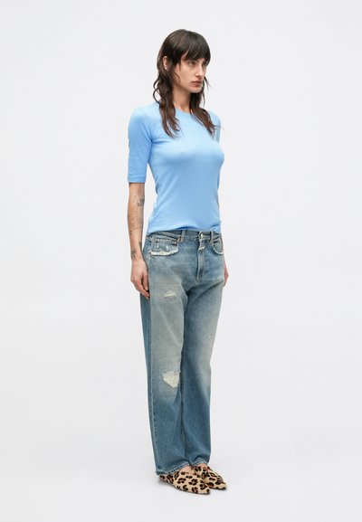 Camicia azzurro chiaro a maniche corte abbinata a jeans azzurri strappati e scarpe slip-on con stampa leopardata, che mostrano uno stile casual.