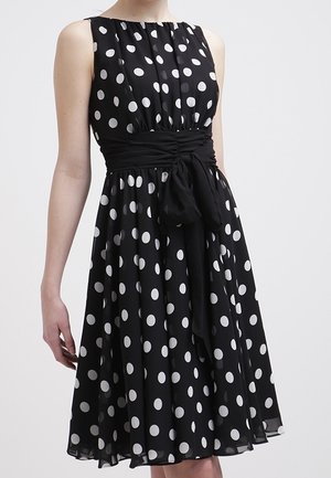 Robe noire sans manches à pois blancs, taille cintrée nouée avec un nœud en tissu, jupe évasée jusqu'aux genoux.