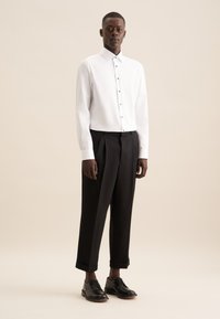 Witte lange kleding met knoopsluiting, gecombineerd met zwarte geplooide broek en zwarte leren schoenen. Eenvoudig ontwerp, gladde textuur.