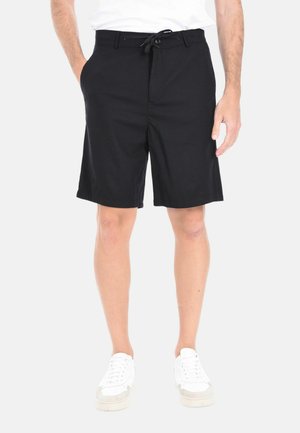 Uomo con pantaloncini neri fino al ginocchio con coulisse e bottone, sneakers bianche e camicia bianca, in piedi con le mani rilassate.