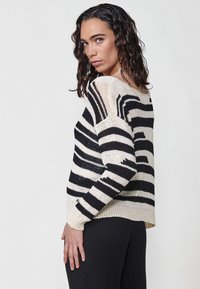 Pulover negru și crem cu dungi, cu o croială lejeră, decolteu larg și manșete cu model împletit. Textură tricotată cu modele de dungi orizontale și verticale.