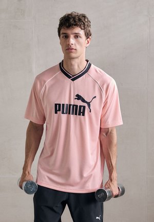 Camisa deportiva de color rosa claro con detalles en negro y el logo de 'PUMA'. Corte holgado, mangas cortas y diseño de cuello en V. Mancuernas grises sujetas con las manos.