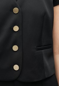 Μαύρο, ραμμένο γιλέκο με τέσσερα μεγάλα στρογγυλά χρυσά κουμπιά και οριζόντια σχισμή τσέπης στη δεξιά πλευρά.