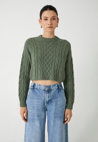 Kurz geschnittenen, grob gestrickten Pullover in Olivgrün mit strukturiertem Zopfmuster und weiten langen Ärmeln, kombiniert mit hellblauen weitem Jeans.