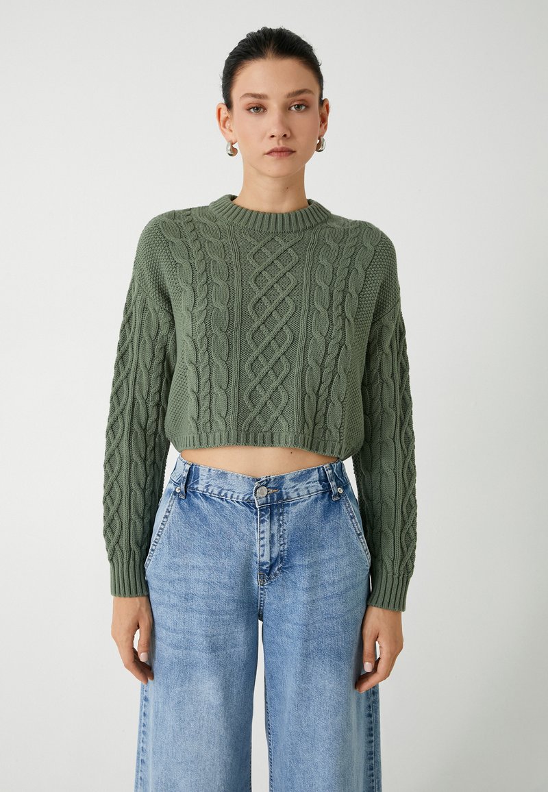 Kurz geschnittenen, grob gestrickten Pullover in Olivgrün mit strukturiertem Zopfmuster und weiten langen Ärmeln, kombiniert mit hellblauen weitem Jeans.