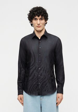 Jonge man met krullend haar, gekleed in een zwart overhemd met patroon en lange mouwen, en lichtblauwe jeans, staand tegen een effen achtergrond.