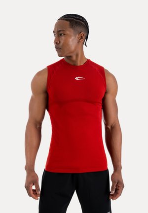 Homme musclé aux cheveux tressés portant un débardeur de sport rouge moulant et un short noir, regardant vers sa gauche sur un fond uni.