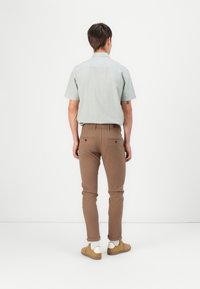 Camisa de manga corta verde claro, pantalones ajustados marrones, puños doblados. Modelo lleva zapatillas color canela con suelas blancas, fondo neutro.