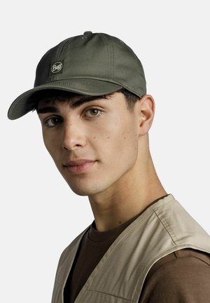 Junger Mann mit grünem Buff-Cap, beiger Weste und braunem Hemd, schaut leicht zur Seite vor einem einfarbigen hellen Hintergrund.