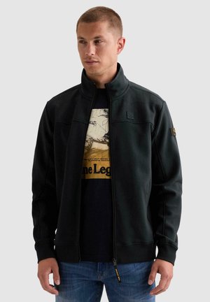 Jeune homme portant une veste noire à zip par-dessus un t-shirt graphique et un jean bleu, regardant légèrement sur le côté sur un fond uni.
