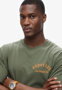 Camiseta de algodón verde oliva con "superdry los ángeles" impreso en naranja. Mangas cortas y cuello redondo. Llevada con un reloj dorado.