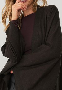 Mørkebrun, blød strikket cardigan med åben frontdesign, draperede ærmer og subtil tekstur. Båret over en maroon-top.