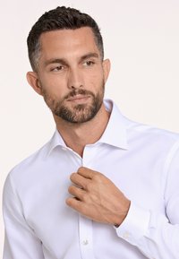 Camisa blanca de vestir con cuello puntiagudo, mangas largas y puños abotonados. Tela suave, brillo sutil y botones en forma de perla.