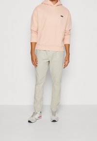 Felpa rosa chiaro con tasca frontale, pantaloni beige e sneakers grigie con strisce bianche e dettagli rossi. Outfit semplice e informale.