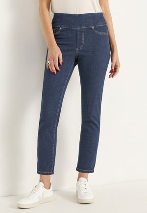 Donkerblauwe spijkerbroek met een brede tailleband, smalle pijpen en decoratieve stiksels. G gedragen met witte sneakers, wat een casual outfit laat zien.