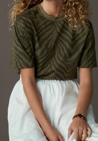 Kobieta w oliwkowozielonej bluzce w zebrę z krótkim rękawem, białej spódnicy z wysokim stanem i czarno-złotym zegarku na nadgarstku, siedząca z rękami opartymi.