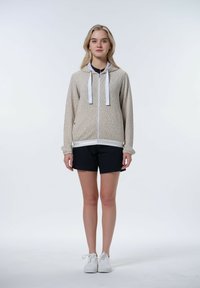 Hellbeige Kapuzenpullover mit Reißverschluss und dezentem Muster, weißen Kordeln und gerippten Bündchen, kombiniert mit marineblauen Shorts und weißen Sneakern.