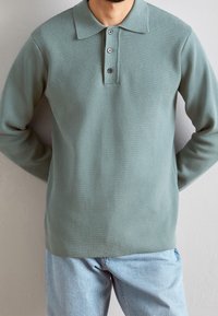 Pull-over vert clair avec un col à revers, une patte à trois boutons et des manches longues. Confectionné en tissu texturé, porté sur un jean bleu clair.